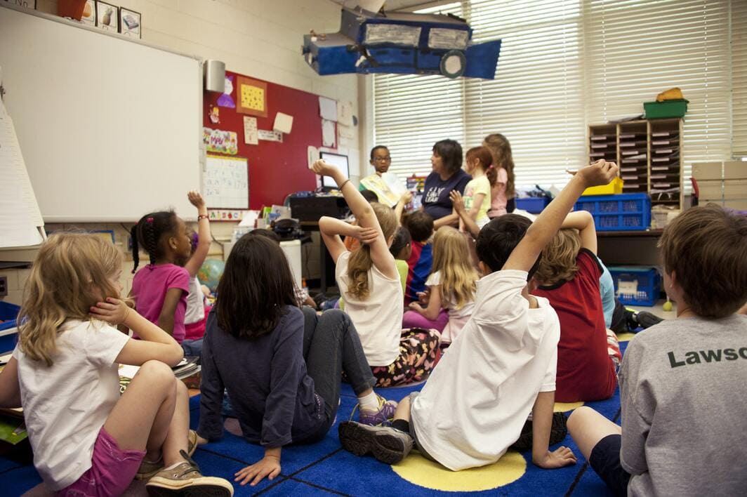 bambini seduti in classe con mani alzate e insegnante sullo sfondo